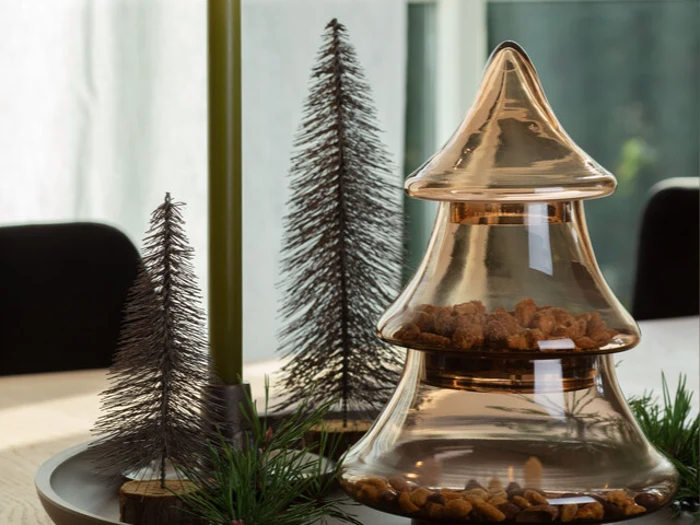 En glasbeholder formet som et juletræ, fyldt med nødder, står på en bakke ved siden af to dekorative metaltræer og grønt løv.
