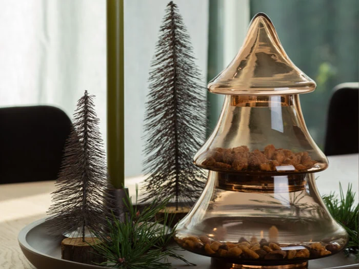 En glaskrukke formet som et juletræ, fyldt med nødder, står på et bord med dekorative metaltræer og grønne fyrretræsgrene.