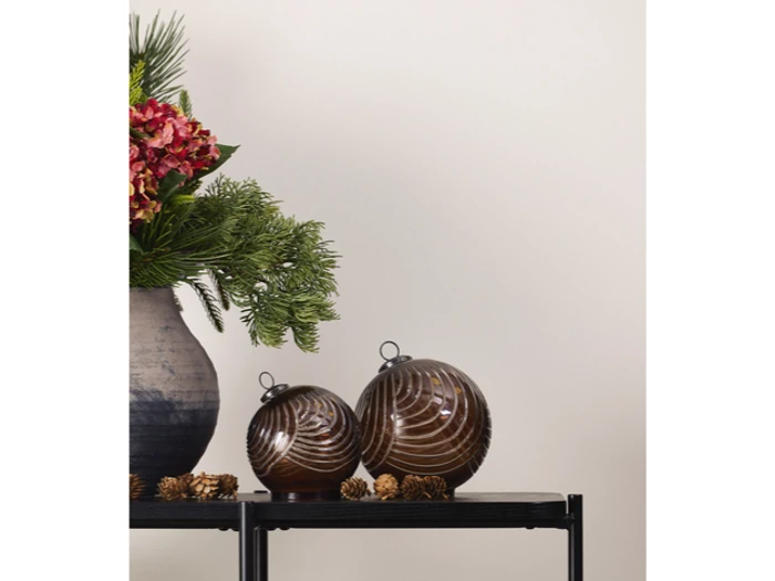 En vase med fyrretræ og røde blomster står ved siden af to runde, brune ornamenter og kogler på et sort bord mod en almindelig, lys væg.