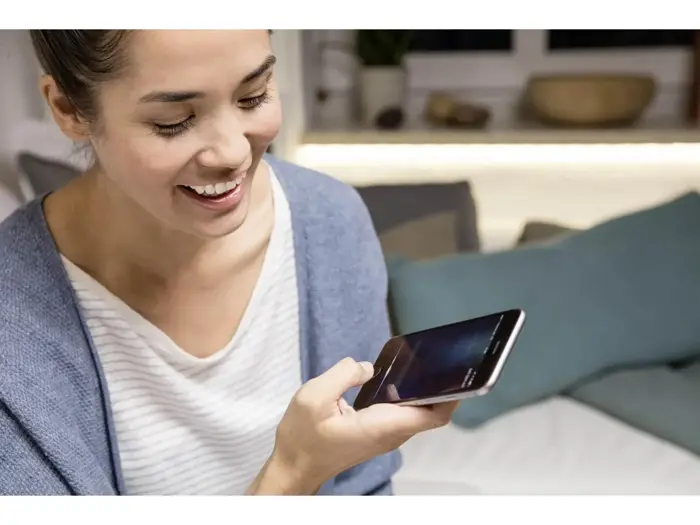 En kvinde smiler, mens hun holder en smartphone og taler ind i den, mens hun sidder indendørs i en sofa med puder i baggrunden.