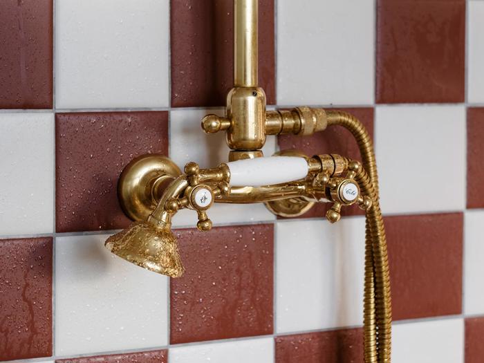 Nærbillede af et messingbrusearmatur i vintagestil med hvide porcelænsdetaljer, monteret på en rød- og hvidternet flisevæg.