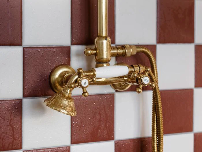 Nærbillede af et messingbrusearmatur i vintagestil med hvide porcelænsdetaljer, monteret på en rød- og hvidternet flisevæg.