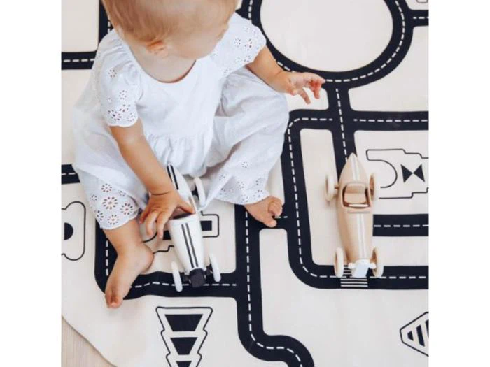 En baby i hvid kjole sidder på en legemåtte med vejmønstre og leger med to legetøjsbiler af træ.