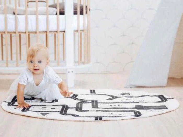 En baby i hvidt tøj kravler på en rund legemåtte med sorte vejmønstre i et lyst børneværelse med en krybbe i baggrunden.