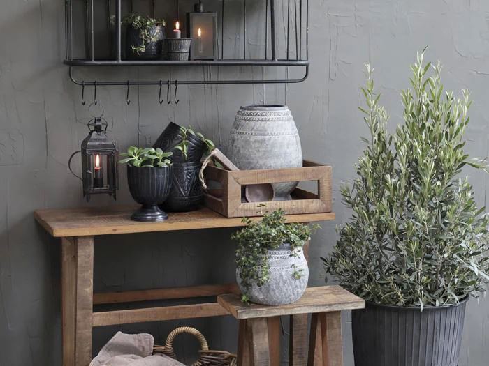 På træborde står potteplanter, vaser og en trækasse ved siden af en bladplante i en sort potte. En hylde og stearinlys hænger på en struktureret grå væg i baggrunden.
