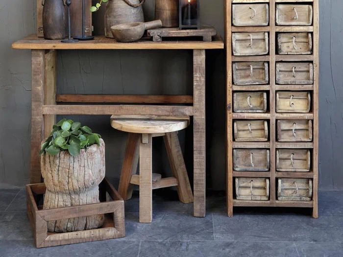Rustikke træmøbler, herunder et bord, en skammel og et skuffeskab, med en potteplante og køkkenudstyr, alt sammen på et gråt stengulv mod en grå væg.