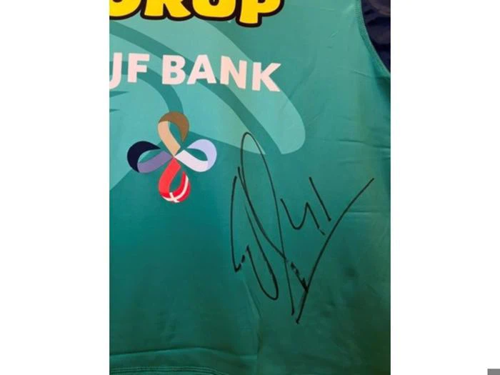 En blågrøn sportstrøje med påskriften JF BANK på forsiden, med et farverigt logo med flere sløjfer og en signatur i sort blæk.