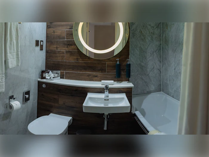 Moderne badeværelse med væghængt håndvask, toilet og badekar. Cirkulært spejl med belysning, træpaneler, marmorvægge og toiletartikler på disken.