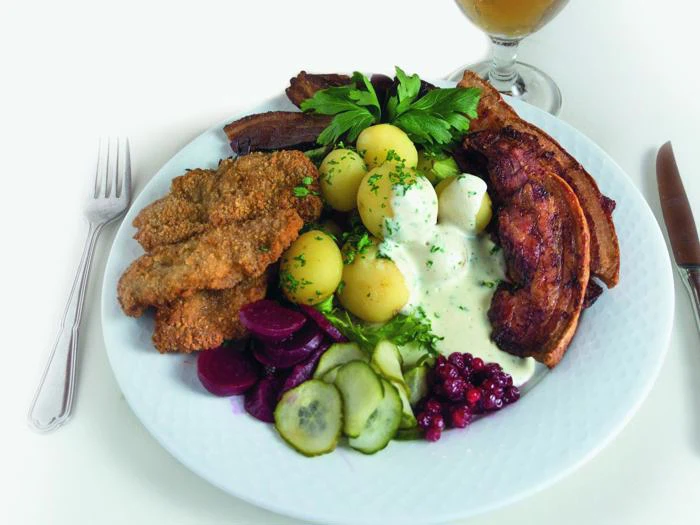 En tallerken med panerede koteletter, stegt flæsk, kogte kartofler med flødesauce, pickles i skiver, rødbeder, tyttebær og frisk persille ved siden af bestik og en drink.