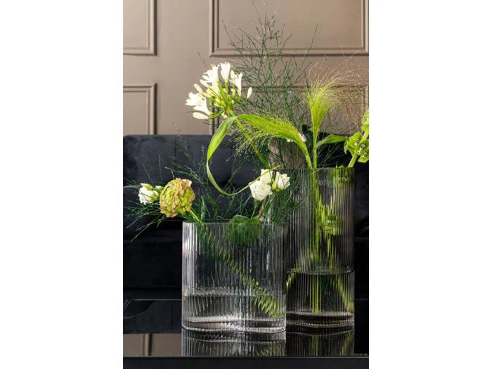 To glasvaser med hvide blomster og grønt løv på en reflekterende sort overflade med en mørk sofa og en panelvæg i baggrunden.