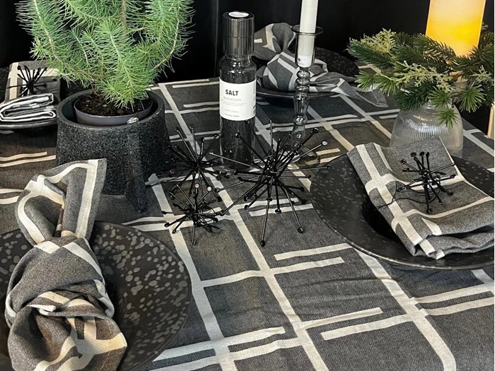 En moderne borddækning med sort og grå geometrisk mønstret dug, sorte tallerkener, foldede servietter, små stedsegrønne potter og en dekorativ saltflaske i midten.