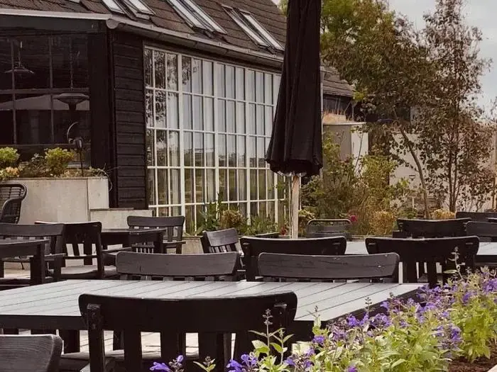 Udendørs siddeområde med sorte borde og stole, lilla blomster i forgrunden og store glasvinduer på en moderne bygning i baggrunden.