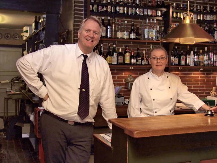 En mand i skjorte og slips og en kvinde i kokkefrakke står bag disken i en hyggelig bar med hylder af flasker bag dem.