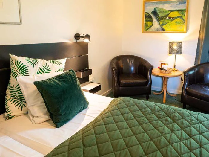 Et hyggeligt hotelværelse med en seng i grønt sengetøj, puder med bladprint og fløjl, to sorte lænestole, et lille rundt bord med en lampe og et landskabsmaleri på væggen.