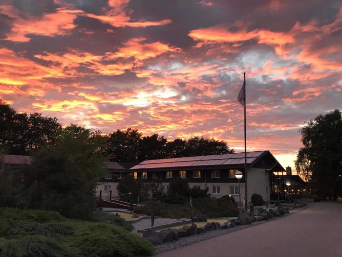 En livlig solnedgang med orange skyer oplyser himlen over en toetagers bygning, træer og en flagstang i et anlagt område.