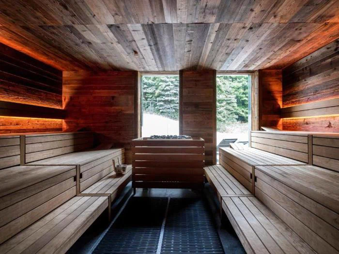 En moderne træsauna med bænke i flere niveauer, varm belysning og store vinduer, der viser en snedækket skov udenfor.