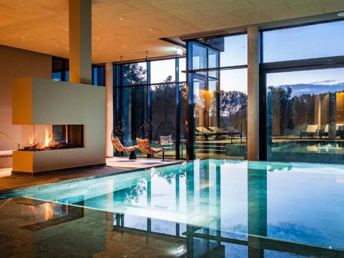 Indendørs poolområde med en moderne pejs, to hængende kurvestole og store gulv-til-loft-vinduer med udsigt til en udendørs scene i skumringen.