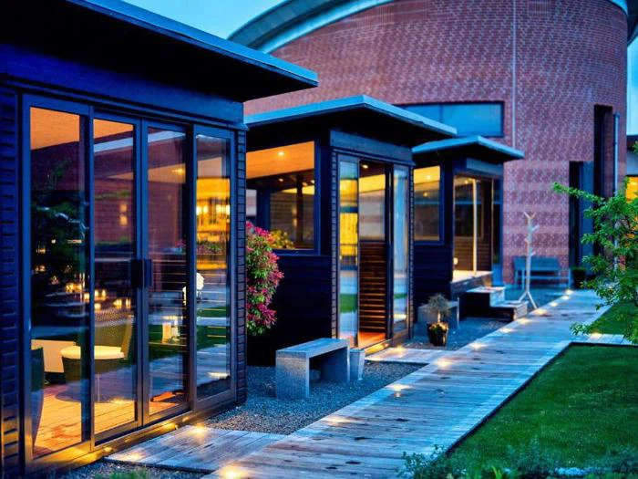 Moderne hytter med glasvægge og varm belysning ligger langs en træsti ved siden af en grøn græsplæne foran en stor murstensbygning i skumringen.