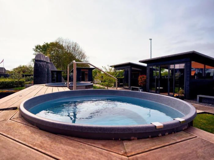 Et rundt udendørs spabad på et trædæk med moderne sorte hytter og grønne områder i baggrunden under en overskyet himmel.