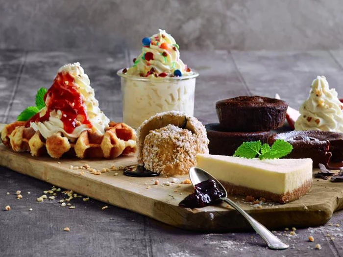 En træplade med forskellige desserter: vafler med fløde og sirup, cheesecake, chokoladekager, is med krymmel og et wienerbrød med flødeskum, pyntet med mynteblade.