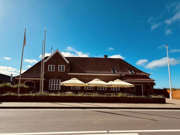 En murstensbygning med rødt tegltag, mærket 1911 over vinduerne, har tre terrasseparasoller udenfor og står under en klar blå himmel med nogle få skyer.