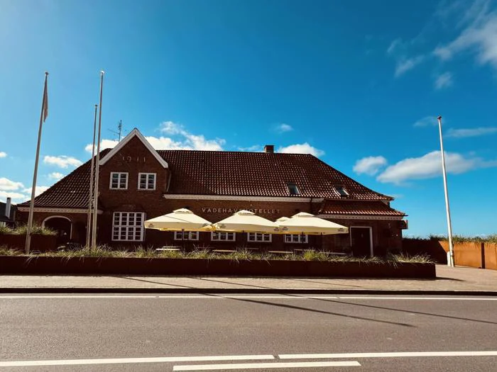 En murstensbygning med rødt tegltag, udendørs parasoller og flag mod en blå himmel med spredte skyer. En vej løber foran bygningen.