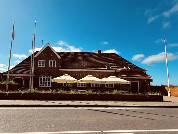 Murstensbygning med rødt tegltag og 1911 markeret på gavlen. Tre gule parasoller skygger for et udendørs siddeområde, og en klar blå himmel er over dem.