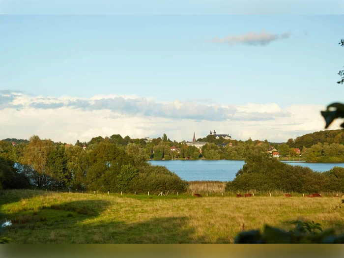 En malerisk udsigt over en sø omgivet af træer og marker, hvor man i det fjerne kan se en by og kirkespir under en klar himmel.