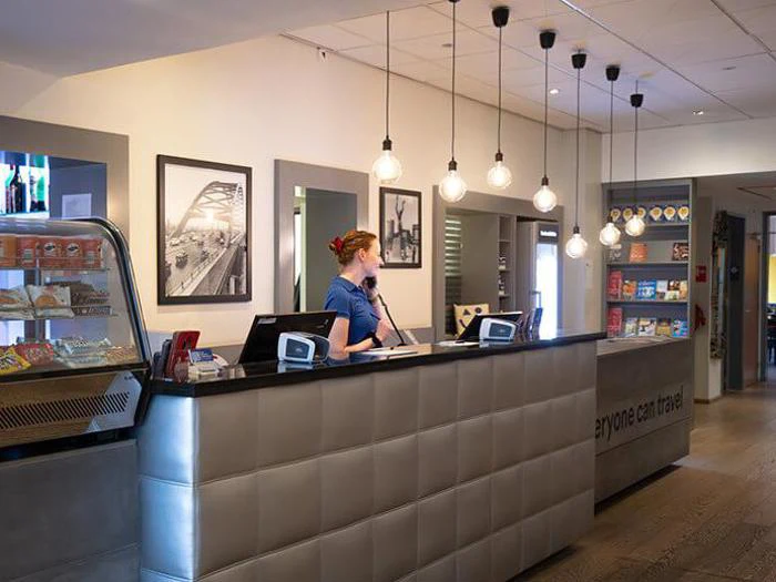 En receptionist står bag en moderne hotelreception og taler i telefon. Hylder med snacks og rejseartikler er synlige i baggrunden sammen med hængelamper og indrammede fotos.