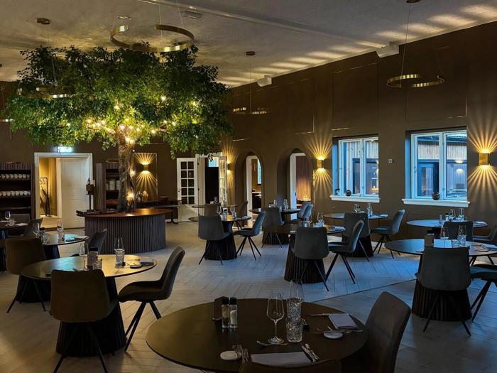 En moderne restaurantspisestue med runde borde, mørke stole, elegant borddækning og et stort træ med lyskæder som midtpunkt, blødt oplyst af varm belysning.