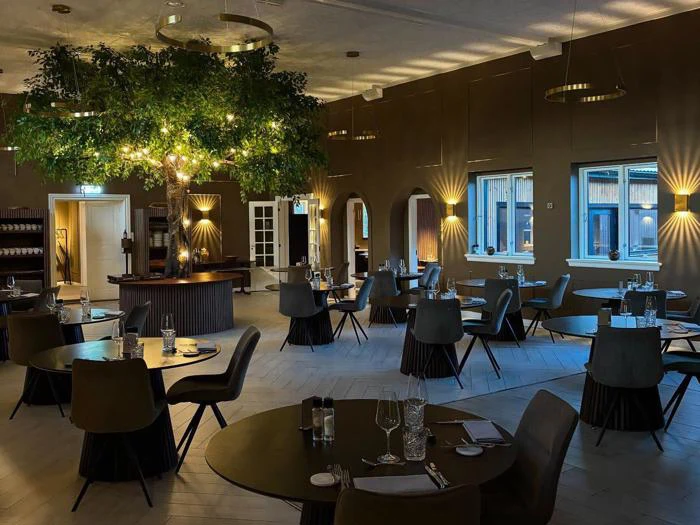 Elegant restaurantinteriør med runde borde, moderne stole, dæmpet belysning og et stort kunstigt træ i midten, som skaber en hyggelig og indbydende spisestemning.