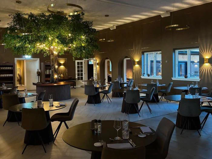 Moderne restaurantspisestue med runde borde, sorte stole, elegant borddækning og et stort kunstigt træ med lys i midten; varm belysning og store vinduer forbedrer stemningen.