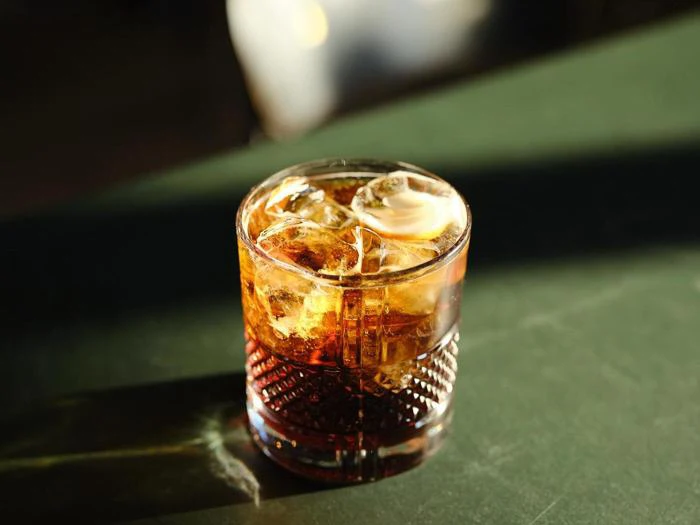 Et glas iskold mørk drik, muligvis whisky eller rom og cola, står på en grøn flade i sollys.