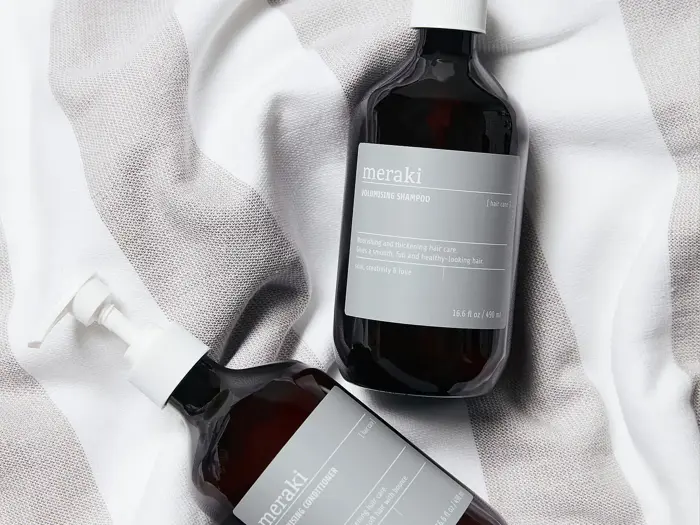To mørke flasker med grå etiketter, en med pumpedispenser og en med hætte, ligger på et hvidt og gråt stribet stof. På etiketterne står der meraki hydrating shampoo og conditioner.