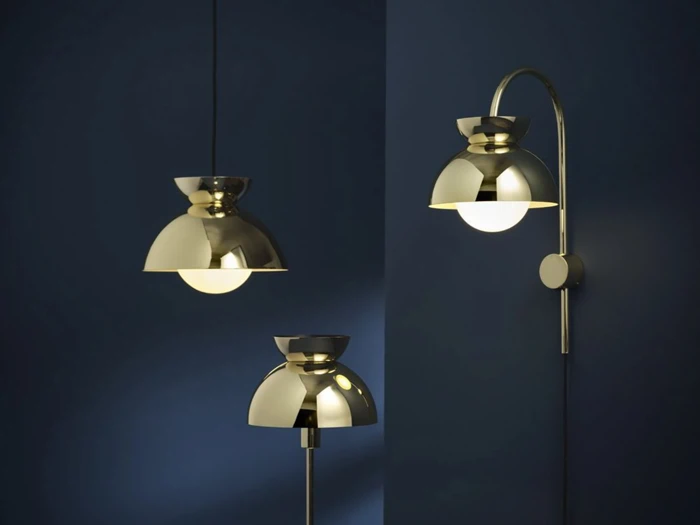 Tre moderne messinglamper med kuppelformede lampeskærme ses foran en mørkeblå væg; én pendel, én gulvlampe og én væglampe er tændt.