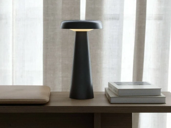 En moderne sort bordlampe står på en træplade ved siden af en lukket laptop og to stablede bøger med beige gardiner i baggrunden.