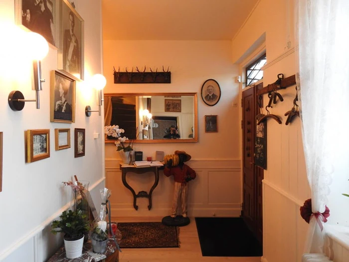 En hyggelig gang med indrammede fotos på væggen, et bord med blomster, et spejl, knager og en lille mannequin klædt i rødt, der står under spejlet.