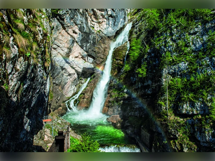 Et vandfald falder ned ad klipperne i en grøn pool med en svag regnbue synlig i tågen og den frodige grønne vegetation, der omgiver scenen.