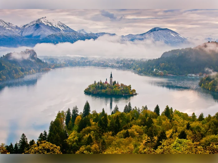 En lille ø med en kirke ligger midt i en sø, omgivet af skovklædte bakker og sneklædte bjerge under en overskyet himmel.