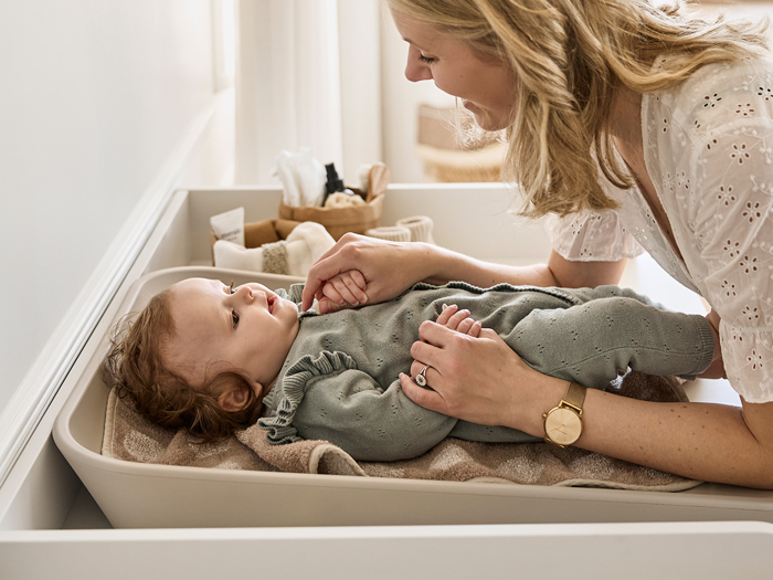 En smilende kvinde holder blidt om en baby, der ligger på et puslebord, og begge har øjenkontakt i et lyst, hyggeligt rum.