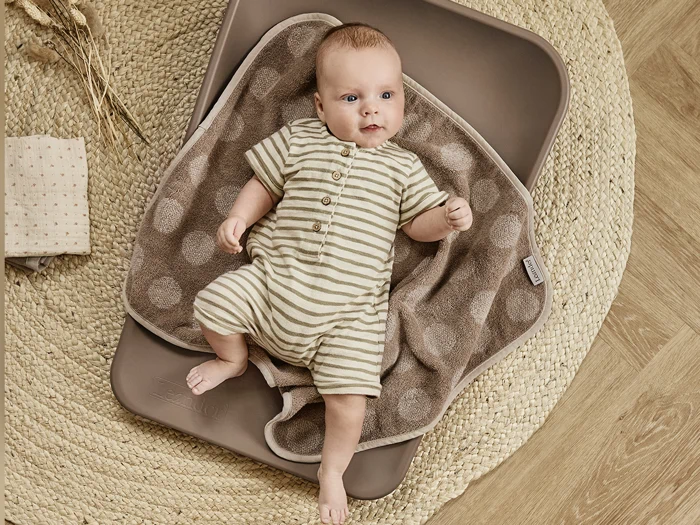 En baby i en stribet sparkedragt ligger på et brunt, prikket håndklæde på en vævet måtte med foldede klude og tørrede blomster i nærheden.