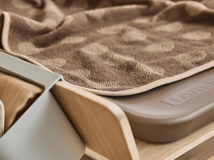 Nærbillede af et brunt tekstureret håndklæde, der er draperet over kanten af en beige puslebakke på en træoverflade med en opbevaringsspand i nærheden.