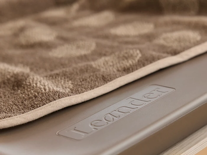 Et nærbillede af et brunt tekstureret håndklæde draperet over en overflade med ordet Leander præget på det.