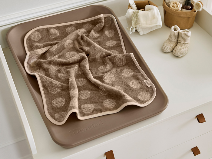 En brun puslepude med et prikket håndklæde, babybooties og små kurve med babyting på en hvid kommode.