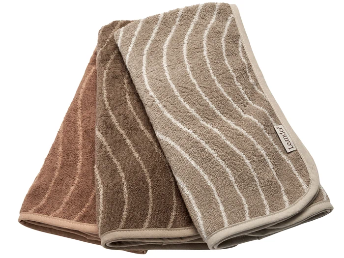 Tre foldede håndklæder i brun, mørkebrun og beige, hver med bølgede hvide linjer og et stofmærke, hvor der står lavendel.