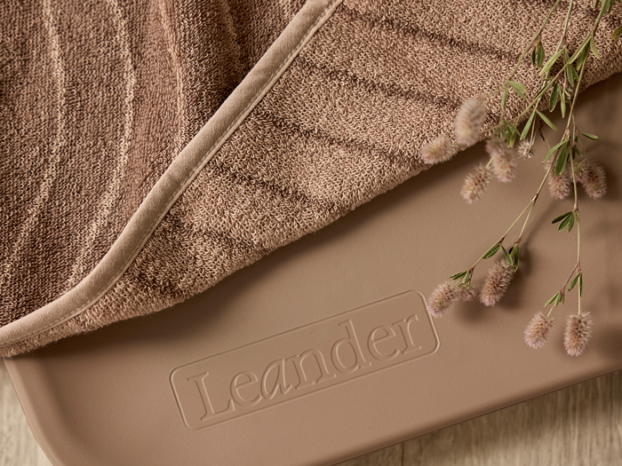 Nærbillede af et blødt brunt håndklæde med en plante, der hviler på en beige overflade med det prægede ord Leander.