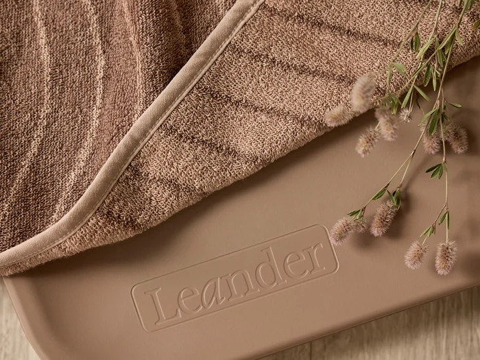 Nærbillede af et blødt brunt håndklæde med en plante, der hviler på en beige overflade med det prægede ord Leander.