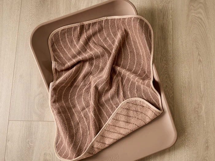 Et brunt, stribet håndklæde er lagt oven på en beige, rektangulær bakke, der står på et lyst trægulv.