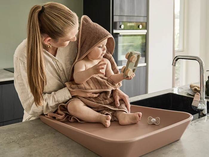 En kvinde holder en baby svøbt i et brunt håndklæde på et køkkenbord. Babyen, som har et håndklæde med hætte på, leger med et stykke trælegetøj, mens en sut ligger i nærheden.