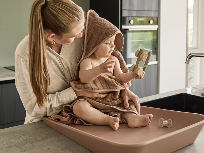 En kvinde holder en baby svøbt i et brunt håndklæde med hætte på en puslepude, mens babyen leger med et stykke trælegetøj. En sut ligger i nærheden på køkkenbordet.